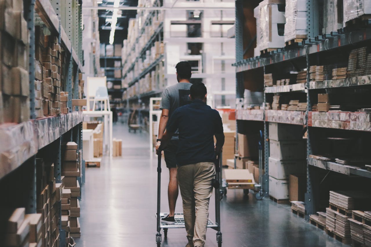 about-01 Two men maneuver a trolley in a large warehouse filled with boxes and shelves.
